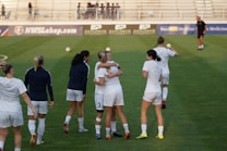 A group of women on a soccer field, some wearing white soccer jerseys and shorts. Two women in the center are embracing, while others are walking or standing nearby. A coach or official can be seen in the distance on the field. The background shows empty bleachers and advertisement banners.
