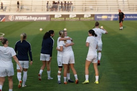 A group of women on a soccer field, some wearing white soccer jerseys and shorts. Two women in the center are embracing, while others are walking or standing nearby. A coach or official can be seen in the distance on the field. The background shows empty bleachers and advertisement banners.