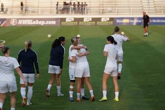 A group of women on a soccer field, some wearing white soccer jerseys and shorts. Two women in the center are embracing, while others are walking or standing nearby. A coach or official can be seen in the distance on the field. The background shows empty bleachers and advertisement banners.