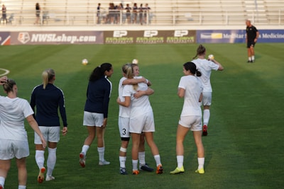 A group of women on a soccer field, some wearing white soccer jerseys and shorts. Two women in the center are embracing, while others are walking or standing nearby. A coach or official can be seen in the distance on the field. The background shows empty bleachers and advertisement banners.