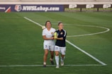 Two female soccer players are walking together on the field, appearing to be in a joyful and celebratory mood. One player is dressed in a white uniform while the other one wears a dark jersey. They are both smiling and holding hands, and the green grass field and a sign for NWSLshop.com are visible in the background.