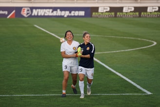 Two female soccer players are walking together on the field, appearing to be in a joyful and celebratory mood. One player is dressed in a white uniform while the other one wears a dark jersey. They are both smiling and holding hands, and the green grass field and a sign for NWSLshop.com are visible in the background.