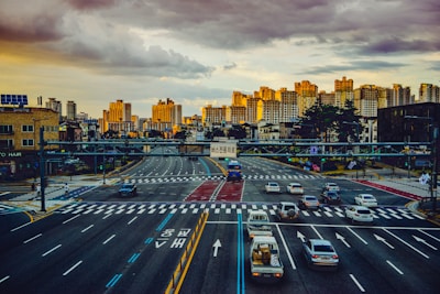 A busy urban intersection with several lanes of traffic, including cars and a bus, under a cloudy sky with golden sunlight illuminating high-rise buildings in the background. Pedestrians can be seen on the sidewalks, and signs with Korean writing are displayed above the road.