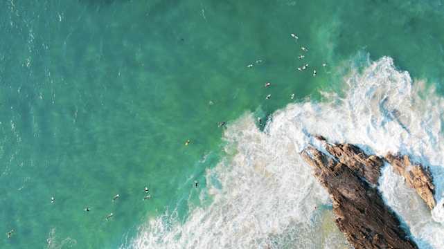 An aerial view of surfers in the ocean near jagged rocks with white waves crashing around them. The water is a striking turquoise color, and surfers are scattered throughout the sea.