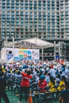 An outdoor event with a large crowd of people seated in plastic chairs. A stage is set up with a large screen displaying colored panels and graphics. Some attendees are wearing shirts labeled 'VOLUNTEER' and colorful clothing, using umbrellas to shield themselves from the sun. The backdrop features a building with a geometric pattern of windows.