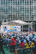 An outdoor event with a large crowd of people seated in plastic chairs. A stage is set up with a large screen displaying colored panels and graphics. Some attendees are wearing shirts labeled 'VOLUNTEER' and colorful clothing, using umbrellas to shield themselves from the sun. The backdrop features a building with a geometric pattern of windows.