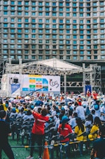 An outdoor event with a large crowd of people seated in plastic chairs. A stage is set up with a large screen displaying colored panels and graphics. Some attendees are wearing shirts labeled 'VOLUNTEER' and colorful clothing, using umbrellas to shield themselves from the sun. The backdrop features a building with a geometric pattern of windows.