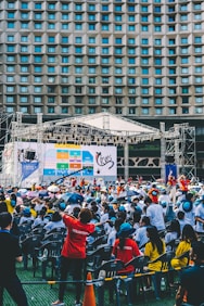 An outdoor event with a large crowd of people seated in plastic chairs. A stage is set up with a large screen displaying colored panels and graphics. Some attendees are wearing shirts labeled 'VOLUNTEER' and colorful clothing, using umbrellas to shield themselves from the sun. The backdrop features a building with a geometric pattern of windows.