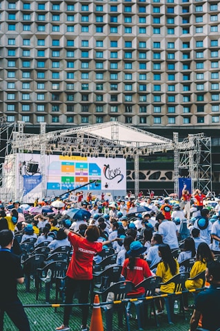 An outdoor event with a large crowd of people seated in plastic chairs. A stage is set up with a large screen displaying colored panels and graphics. Some attendees are wearing shirts labeled 'VOLUNTEER' and colorful clothing, using umbrellas to shield themselves from the sun. The backdrop features a building with a geometric pattern of windows.