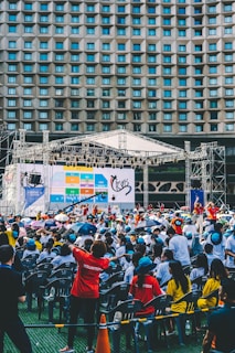 An outdoor event with a large crowd of people seated in plastic chairs. A stage is set up with a large screen displaying colored panels and graphics. Some attendees are wearing shirts labeled 'VOLUNTEER' and colorful clothing, using umbrellas to shield themselves from the sun. The backdrop features a building with a geometric pattern of windows.