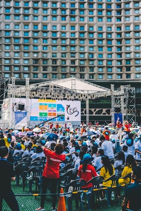 An outdoor event with a large crowd of people seated in plastic chairs. A stage is set up with a large screen displaying colored panels and graphics. Some attendees are wearing shirts labeled 'VOLUNTEER' and colorful clothing, using umbrellas to shield themselves from the sun. The backdrop features a building with a geometric pattern of windows.