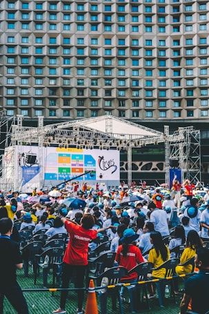 An outdoor event with a large crowd of people seated in plastic chairs. A stage is set up with a large screen displaying colored panels and graphics. Some attendees are wearing shirts labeled 'VOLUNTEER' and colorful clothing, using umbrellas to shield themselves from the sun. The backdrop features a building with a geometric pattern of windows.