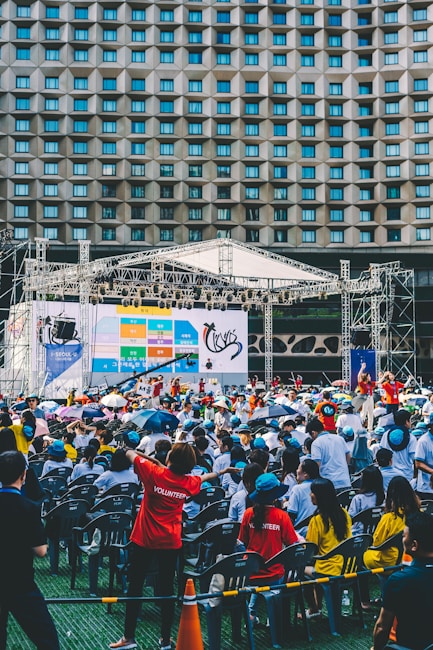 An outdoor event with a large crowd of people seated in plastic chairs. A stage is set up with a large screen displaying colored panels and graphics. Some attendees are wearing shirts labeled 'VOLUNTEER' and colorful clothing, using umbrellas to shield themselves from the sun. The backdrop features a building with a geometric pattern of windows.
