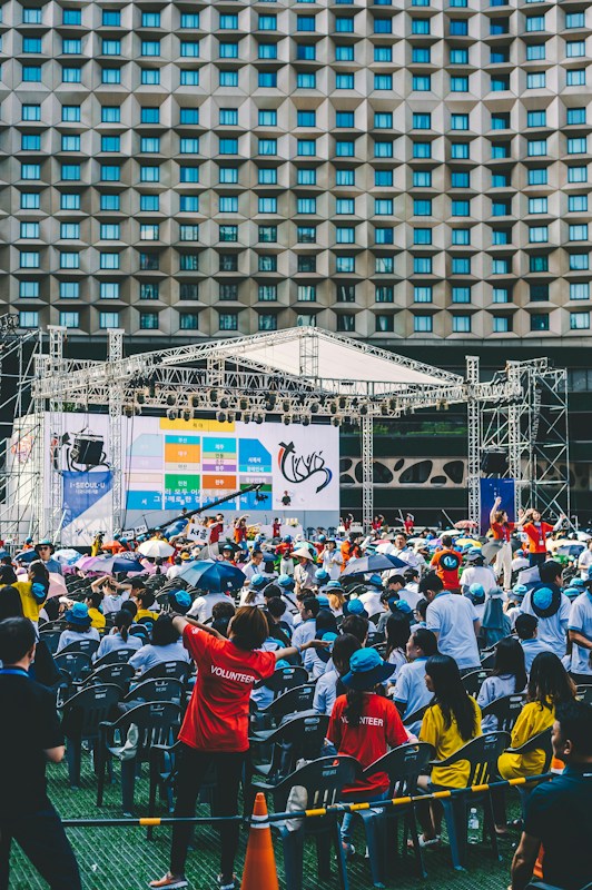 An outdoor event with a large crowd of people seated in plastic chairs. A stage is set up with a large screen displaying colored panels and graphics. Some attendees are wearing shirts labeled 'VOLUNTEER' and colorful clothing, using umbrellas to shield themselves from the sun. The backdrop features a building with a geometric pattern of windows.
