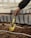 A hand holding a small, yellow gardening trowel digging into soil. The trowel has visible wear and discoloration from use. The background shows soft-focus raised garden beds with soil and protective covers or barriers.