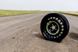 A black tire with visible green labels is positioned on a dirt road. The road stretches into the distance with grassy fields on either side, under a light, overcast sky.