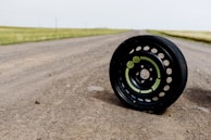 A black tire with visible green labels is positioned on a dirt road. The road stretches into the distance with grassy fields on either side, under a light, overcast sky.
