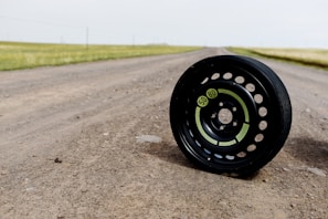 A black tire with visible green labels is positioned on a dirt road. The road stretches into the distance with grassy fields on either side, under a light, overcast sky.