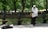A musician is playing a saxophone in a park setting, wearing a white shirt and cap. The background features lush green trees and benches, and an instrument case is lying on the ground nearby.