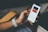 A person holds a smartphone displaying a music app interface. The screen shows a song titled 'Furious' by Jade Bird. The individual's hand is adorned with a white perforated watch band. The background includes a wooden armrest and a blurred pair of shorts.