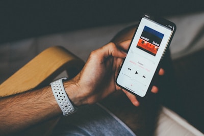 A person holds a smartphone displaying a music app interface. The screen shows a song titled 'Furious' by Jade Bird. The individual's hand is adorned with a white perforated watch band. The background includes a wooden armrest and a blurred pair of shorts.