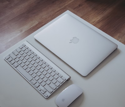 A closed silver laptop with an Apple logo is placed on a white desk. Next to it, there is a white wireless keyboard and a white wireless mouse. The surface of the desk is clean and the background shows a wooden floor.