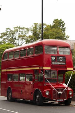 A classic red double-decker bus with signage for a wedding party is parked on a street. The bus is decorated with ribbons and has a sign that reads '2018 Happy Wedding Day' above the windshield. It is surrounded by greenery and trees in the background.