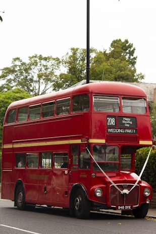 A classic red double-decker bus with signage for a wedding party is parked on a street. The bus is decorated with ribbons and has a sign that reads '2018 Happy Wedding Day' above the windshield. It is surrounded by greenery and trees in the background.