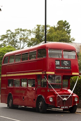 A classic red double-decker bus with signage for a wedding party is parked on a street. The bus is decorated with ribbons and has a sign that reads '2018 Happy Wedding Day' above the windshield. It is surrounded by greenery and trees in the background.