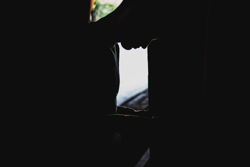 A silhouette of a person in a contemplative posture is captured in low lighting, with a bright background contrast that highlights their shape. The scene conveys solitude and introspection.