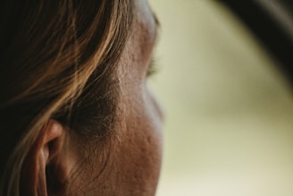 A close-up of a person's profile, focusing on the ear and side of the face. The background is blurred, suggesting a shallow depth of field.