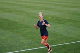 A person is jogging on a grassy field while wearing a navy blue shirt with a logo and red shorts. They are making a hand gesture and appear focused.