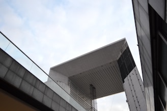 A striking modern architectural structure with geometric lines and a large, cubic shape. It features gray and black metal panels, creating a sleek and industrial look. The structure is surrounded by glass railings and another building partially visible in the foreground.