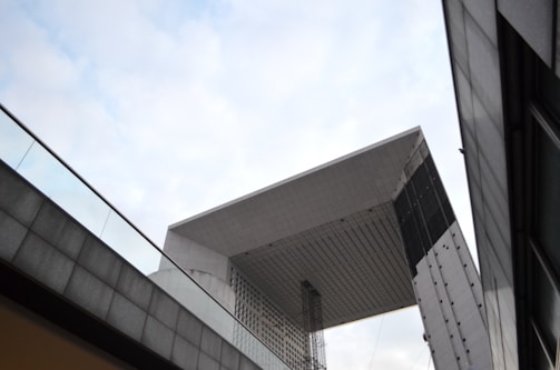 A striking modern architectural structure with geometric lines and a large, cubic shape. It features gray and black metal panels, creating a sleek and industrial look. The structure is surrounded by glass railings and another building partially visible in the foreground.