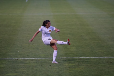 A female soccer player in a white uniform is kicking a ball on a green soccer field. The player has dark hair tied back and is focused on the action. The uniform has a prominent logo, and the player is wearing white socks and cleats with red accents.