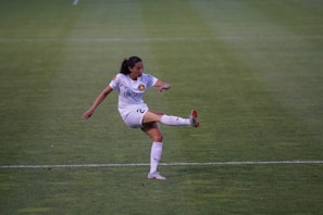 A female soccer player in a white uniform is kicking a ball on a green soccer field. The player has dark hair tied back and is focused on the action. The uniform has a prominent logo, and the player is wearing white socks and cleats with red accents.