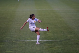 A female soccer player in a white uniform is kicking a ball on a green soccer field. The player has dark hair tied back and is focused on the action. The uniform has a prominent logo, and the player is wearing white socks and cleats with red accents.