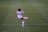 A female soccer player in a white uniform is kicking a ball on a green soccer field. The player has dark hair tied back and is focused on the action. The uniform has a prominent logo, and the player is wearing white socks and cleats with red accents.