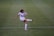 A female soccer player in a white uniform is kicking a ball on a green soccer field. The player has dark hair tied back and is focused on the action. The uniform has a prominent logo, and the player is wearing white socks and cleats with red accents.