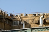 A row of air conditioning units is installed on the rooftop of a building. A person, wearing a hat, is working on one of the units. The building has several ventilators and metal ducts extending from it. The sky is clear and blue.