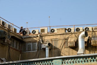 A row of air conditioning units is installed on the rooftop of a building. A person, wearing a hat, is working on one of the units. The building has several ventilators and metal ducts extending from it. The sky is clear and blue.