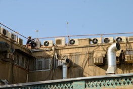 A row of air conditioning units is installed on the rooftop of a building. A person, wearing a hat, is working on one of the units. The building has several ventilators and metal ducts extending from it. The sky is clear and blue.