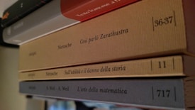 Four books are stacked horizontally on a shelf. The spines of the books are visible, displaying titles written in Italian. The titles read, from top to bottom: 'Testo francese al...' (partial), 'Così parlò Zarathustra', 'Sull'utilità e il danno della storia', and 'L'arte della matematica'. The covers are in different shades, including red, beige, and gray.