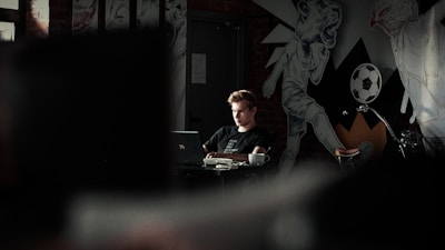 A person is seated at a table with a laptop, surrounded by eclectic murals and artwork on the walls. The environment appears to be a café or creative workspace, with dim lighting and a moody atmosphere. A coffee cup sits on the table next to the person.