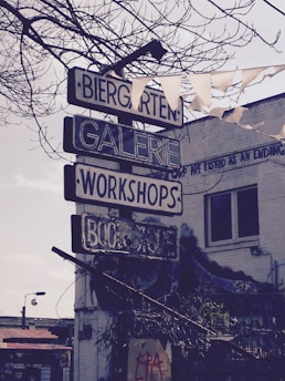 A vintage-style signpost with multiple directional signs pointing to a Biergarten, Galerie, Workshops, and Bookstore. These signs are mounted on a pole in a rustic outdoor setting with an overcast sky and bare tree branches partially visible. A partially visible building shows urban graffiti and art on its brick facade.