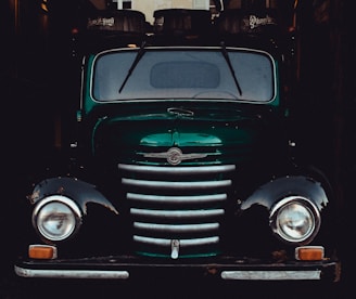 A vintage, green truck with a distinctive front grille and round headlights. It is parked in a dimly lit space, with barrels labeled 'Pilsner Urquell' visible on top, likely loaded for transport.