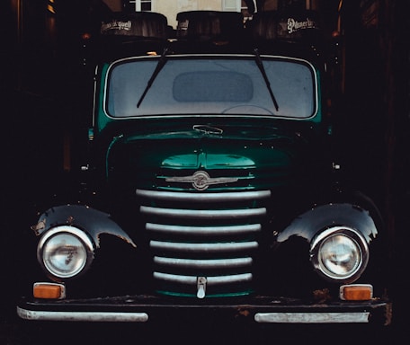 A vintage, green truck with a distinctive front grille and round headlights. It is parked in a dimly lit space, with barrels labeled 'Pilsner Urquell' visible on top, likely loaded for transport.