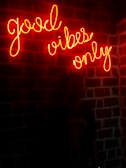 A neon sign with the words 'good vibes only' is mounted on a brick wall. The glowing text casts a warm, red-orange hue over the wall, creating an inviting and energetic atmosphere. A silhouette of a person is faintly visible standing in front of the wall, adding a subtle human element to the scene.