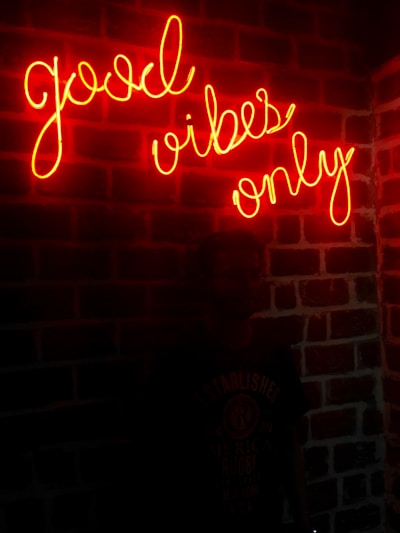 A neon sign with the words 'good vibes only' is mounted on a brick wall. The glowing text casts a warm, red-orange hue over the wall, creating an inviting and energetic atmosphere. A silhouette of a person is faintly visible standing in front of the wall, adding a subtle human element to the scene.