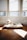 A small, fluffy dog is lying on a cushioned pet bed placed on a wooden floor. The space is brightly lit by natural sunlight coming through three windows with partially closed blinds.